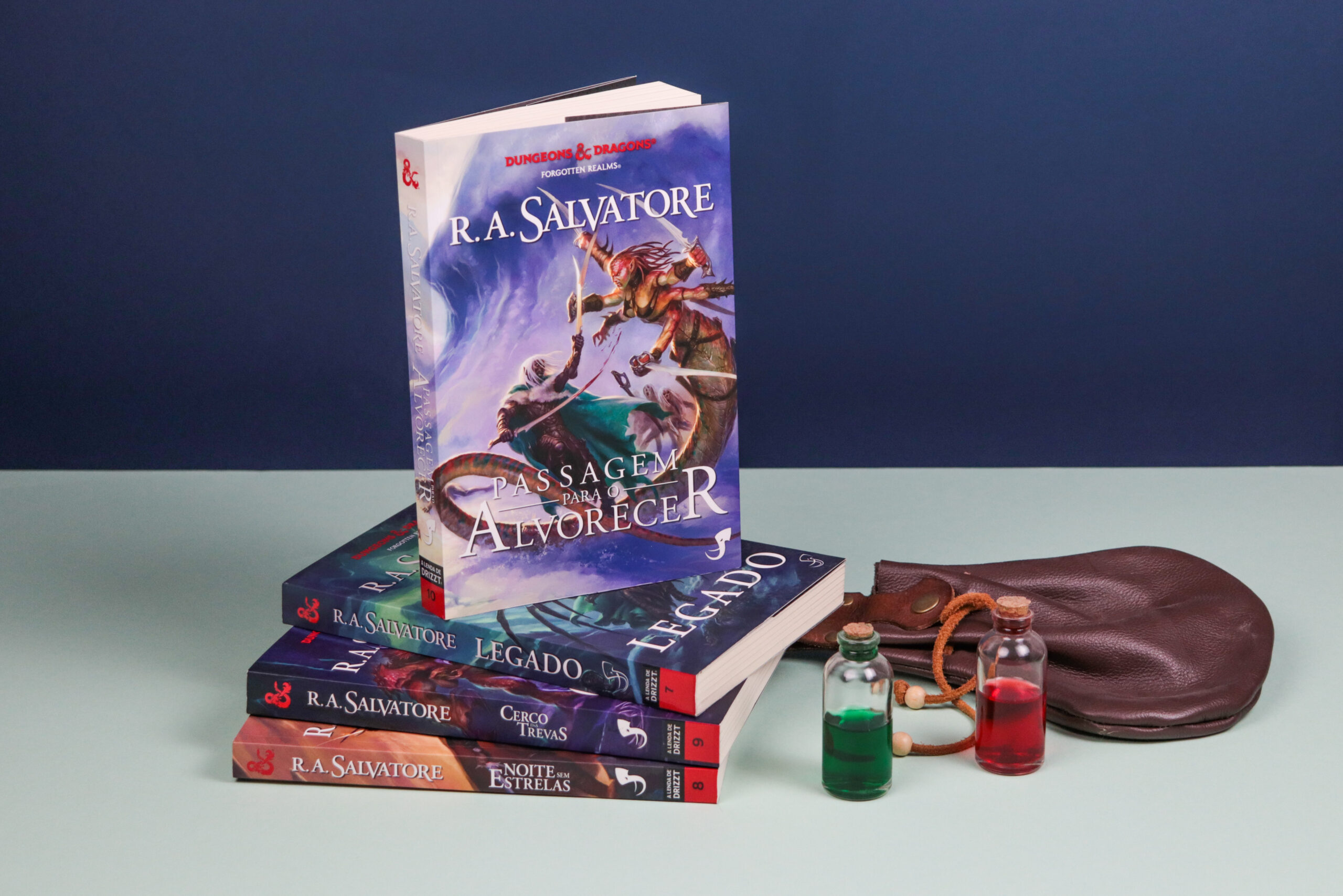 Livro Passagem para o Alvorecer em pé, em cima de outros livros da série Drizzt. Na direita dois pequenos frascos de vidro com líquido vermelho e verde e uma algibeira de couro.